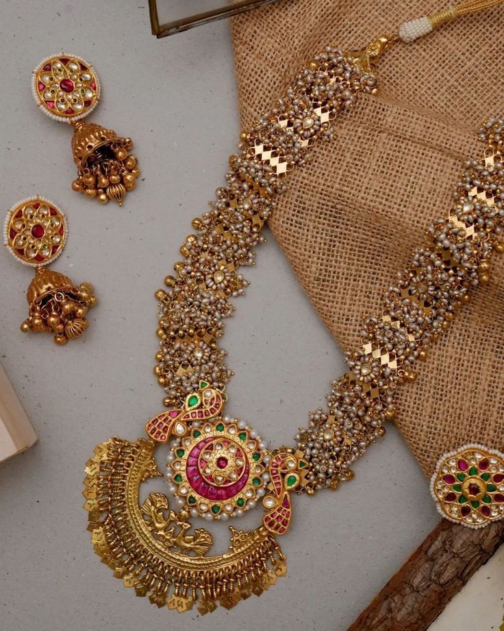 Gold necklace with intricate designs and red and green stones on a textured surface.