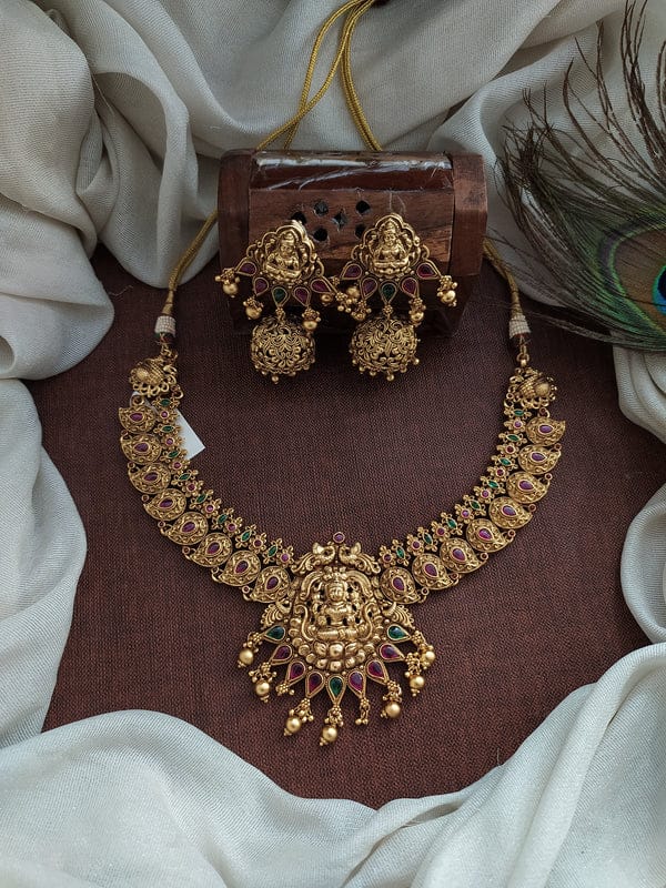 Gold necklace and earrings set on a brown surface with white fabric and peacock feather in the background