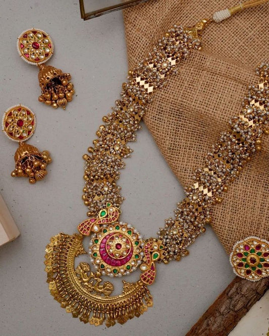 Gold necklace with intricate designs and red and green stones on a textured surface.
